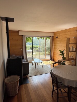 A cozy room with a wooden interior, a large window, a sofa, a dining table, and a small fireplace. - Charming & Cozy 2-Bedroom Apartment in Talloires: Garden and Lake View (Talloires-Montmin)