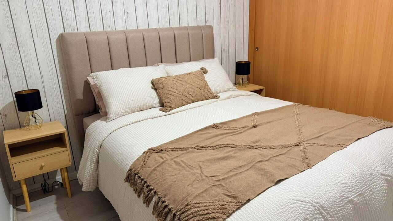 A neatly made bed with a beige headboard, two bedside tables with lamps, and a wooden door in the background.