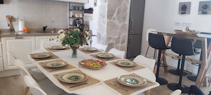 A dining area with a white table set for six, surrounded by white chairs. A coffee machine and a refrigerator are visible in the background. - Mama's House, where tradition and comfort will make you feel right at home (Mijas)