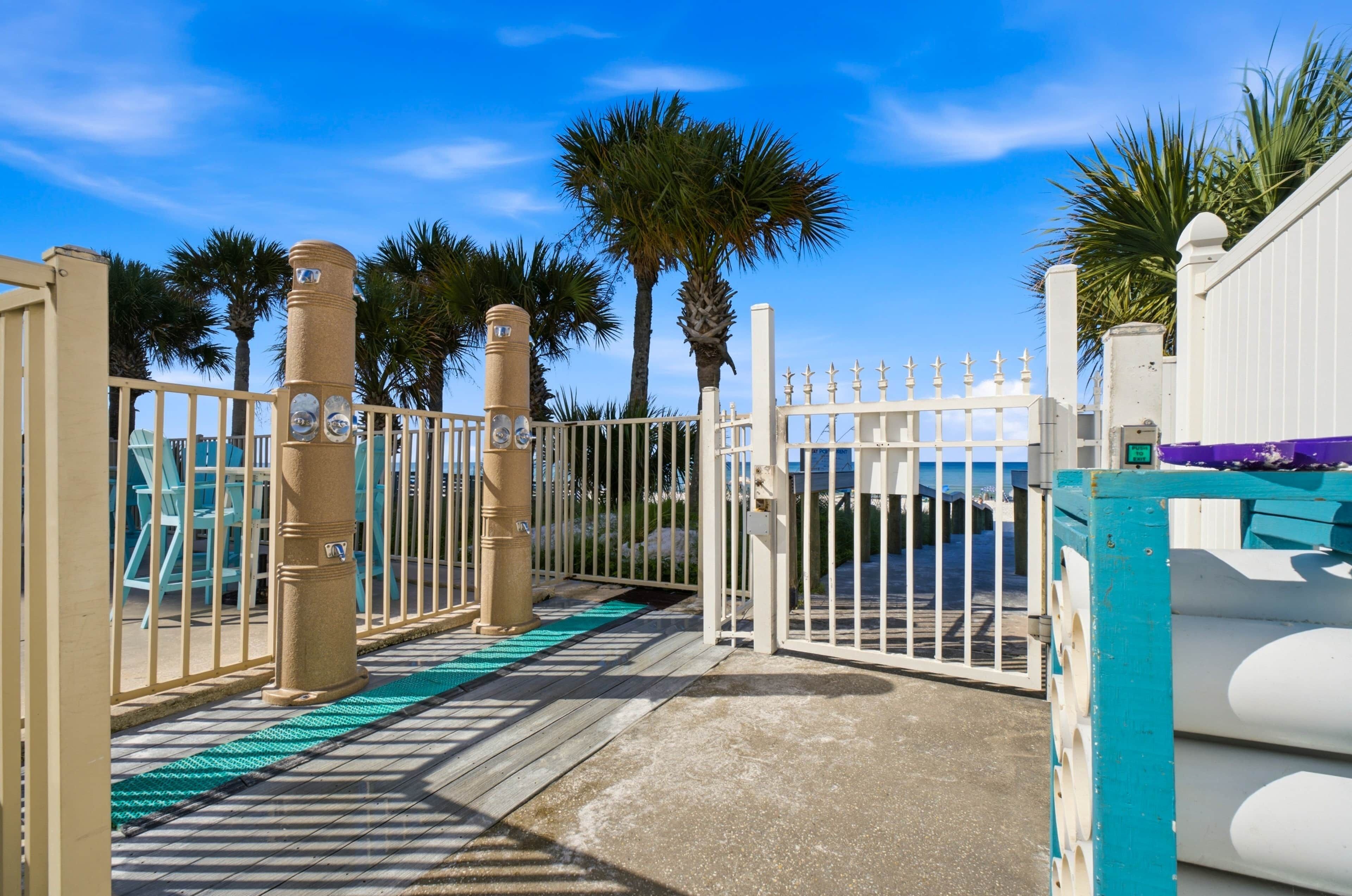 A gated entrance with a wooden deck leading to a beach area, palm trees, and a clear blue sky.