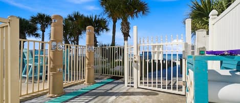 Una entrada con reja y una terraza de madera que conduce a una zona de playa, palmeras y un cielo azul despejado.