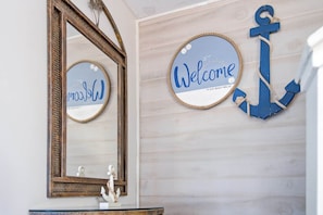 A bathroom with a large mirror, a welcome sign, and a decorative anchor. - Gulf Highlands with a Golf Cart! (Panama City Beach)
