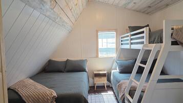 Une chambre mansardée avec un plafond en pente, deux lits superposés, une petite fenêtre et un plancher en bois.