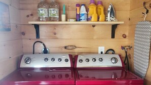 A compact kitchen with a red stove and oven, a wooden shelf above holding various kitchen items, and a patterned towel hanging on the wall.