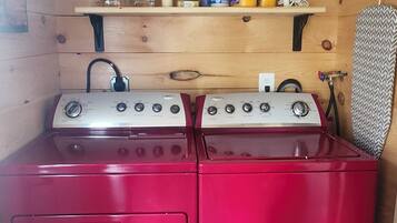 Une cuisine compacte avec un poêle et un four rouges, une étagère en bois au-dessus où sont rangés divers ustensiles de cuisine, et une serviette à motifs accrochée au mur.
