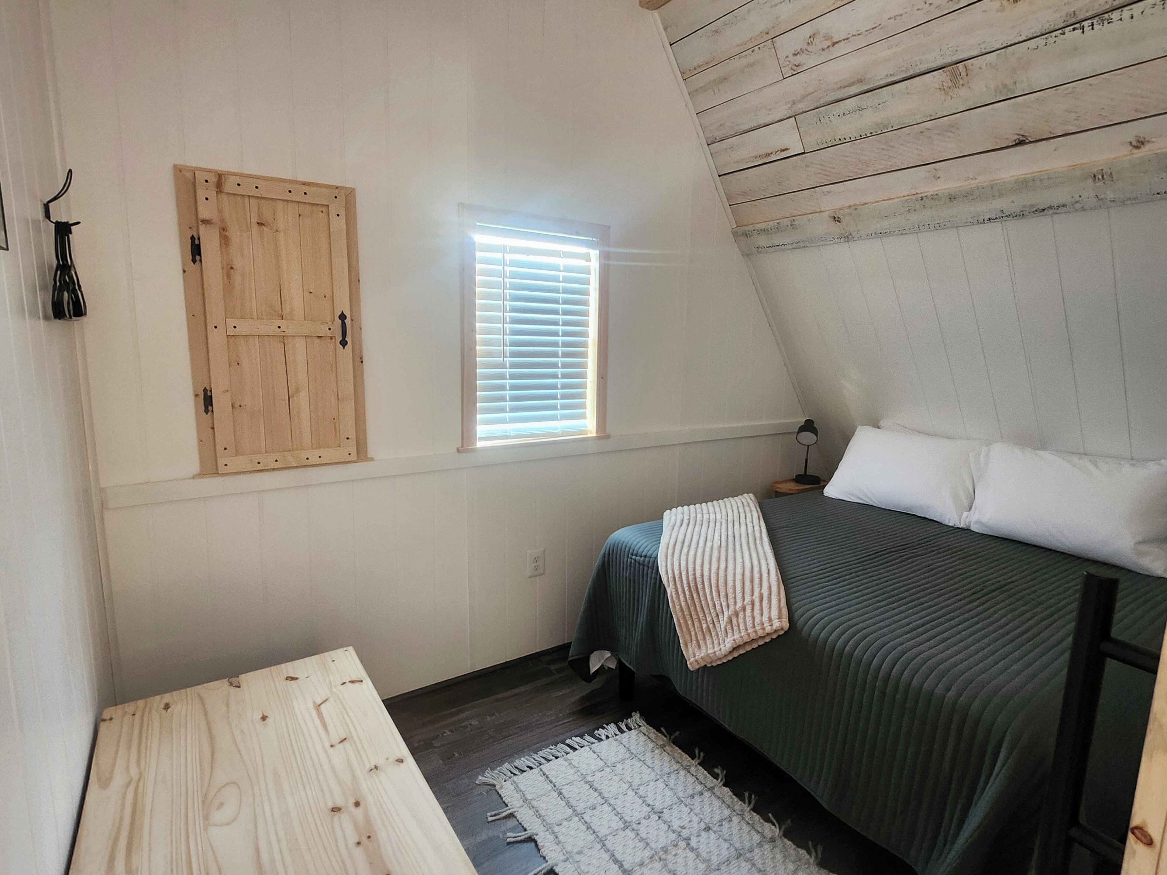 Ein Schlafzimmer mit schrägverdeckter Decke, einem Bett mit Kissen, einem Nachttisch, einer Lampe, einem Fenster mit Jalousien und einer Holztür.