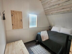 A bedroom with a sloped ceiling, a bed with pillows, a bedside table, a lamp, a window with blinds, and a wooden door.