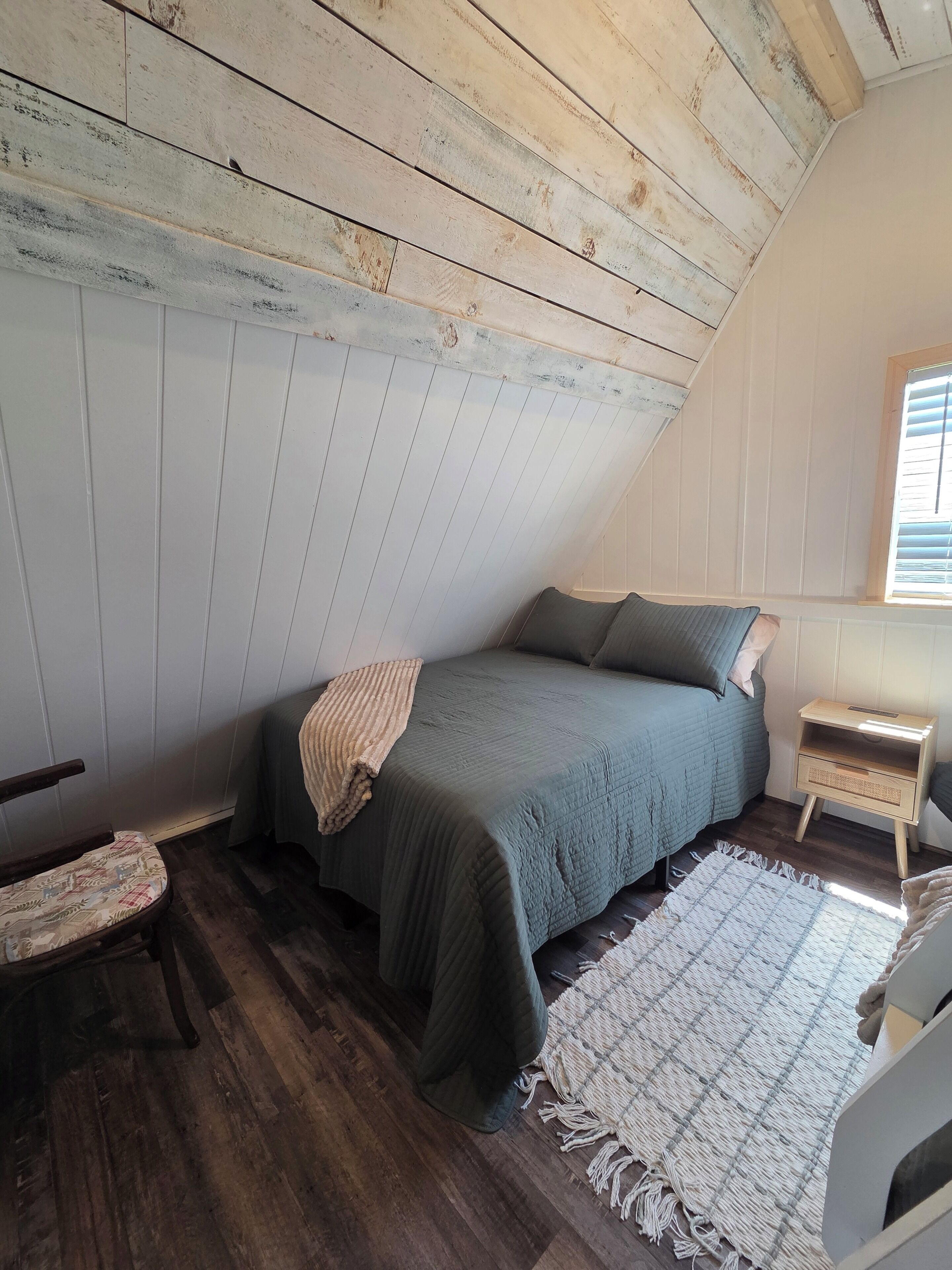 A bedroom with a sloped ceiling, a bed with a green bedspread, a chair with a patterned cushion, a small bedside table, and a window with blinds.