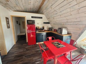 A compact kitchen with a red refrigerator, a microwave, and a toaster. There is a dining table with red chairs and a small rug.