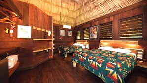 A room with wooden walls and a thatched roof, featuring two beds with colorful blankets, a small table, and a shelf with a fire extinguisher.