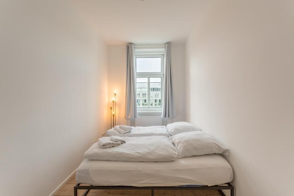 Een eenpersoonsbed met witte beddengoed in een kamer met een raam en een bedlampje.