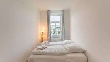 Een eenpersoonsbed met witte beddengoed in een kamer met een raam en een bedlampje.