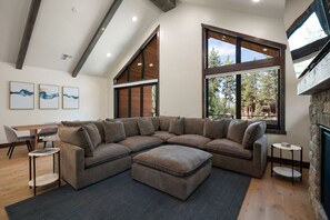 A modern living room with a sectional sofa, a dining table, and large windows offering a view of the outdoors. - Sierra Luxe Retreat - Hot tub close to Stateline (Zephyr Cove)