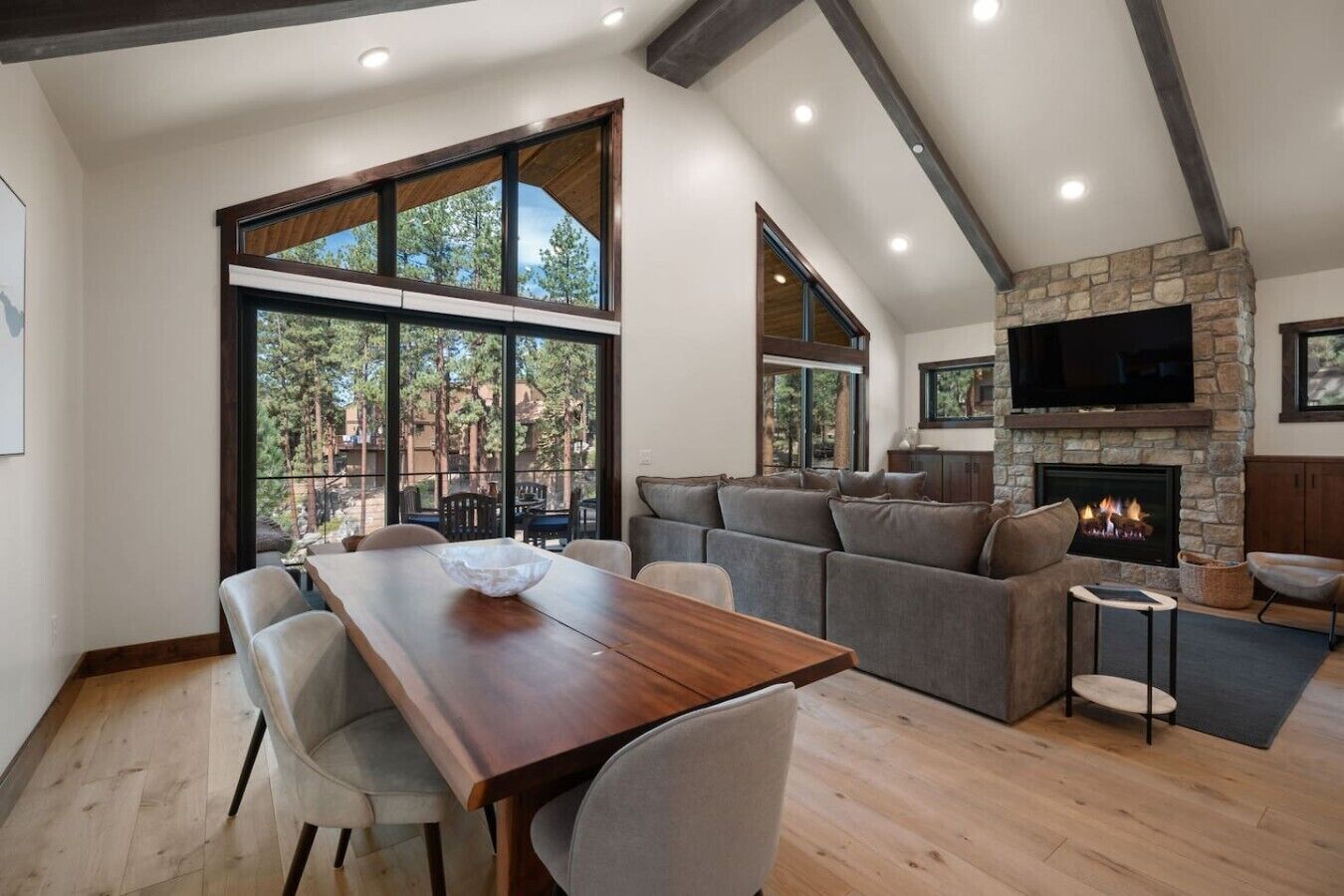 A modern living room with a stone fireplace, a large TV mounted above it, and a dining area with a wooden table and chairs.