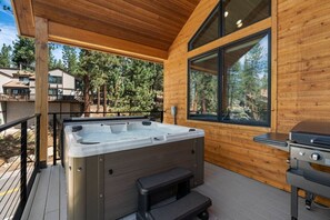 Una terraza de madera con un jacuzzi, una parrilla y un ventanal grande con vista a un bosque.