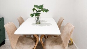 Una mesa blanca con un jarrón de vidrio transparente que contiene una planta, rodeada por cuatro sillas color beige con patas de madera.