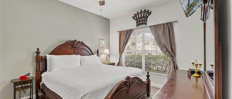 A bedroom with a wooden bed, a nightstand, a bedside table, a ceiling fan, a window with blinds, a mirror, and a wall-mounted TV.