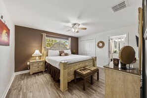 Ein Schlafzimmer mit einem Bett, Nachttischen, einem Deckenventilator, einem Fenster mit Vorhängen, einem Spiegel und einer Wanddekoration.