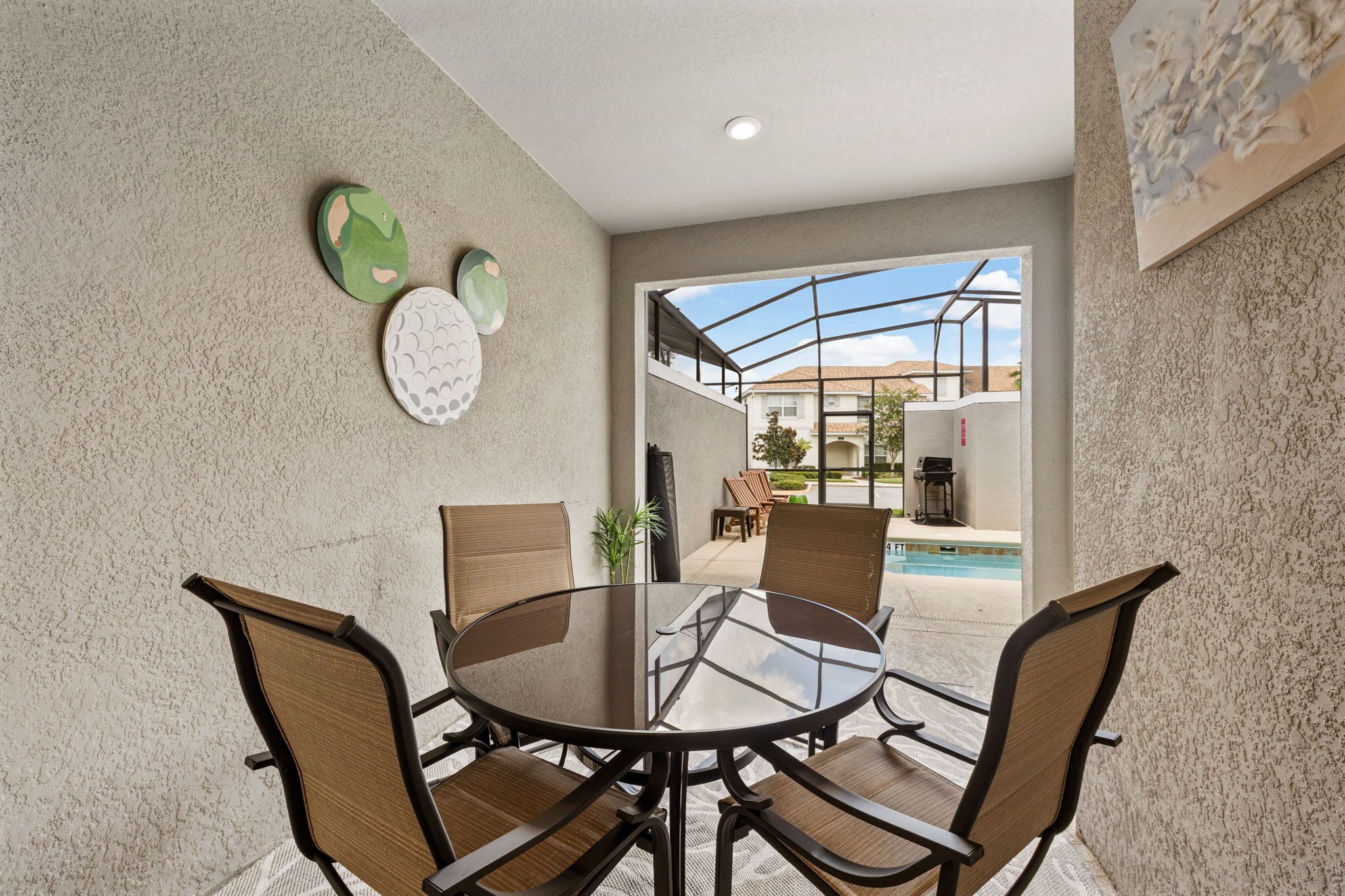 A dining area with a round glass table and four chairs, a pool in the background, and a view of a residential area.