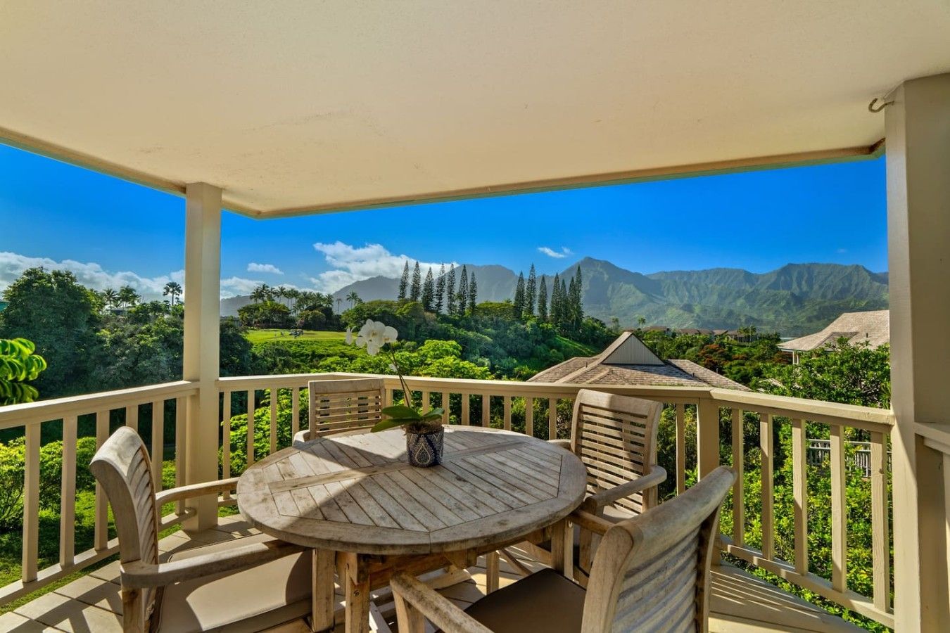 Um deck de madeira com uma mesa redonda e cadeiras, com vista para uma paisagem exuberante com montanhas e árvores.