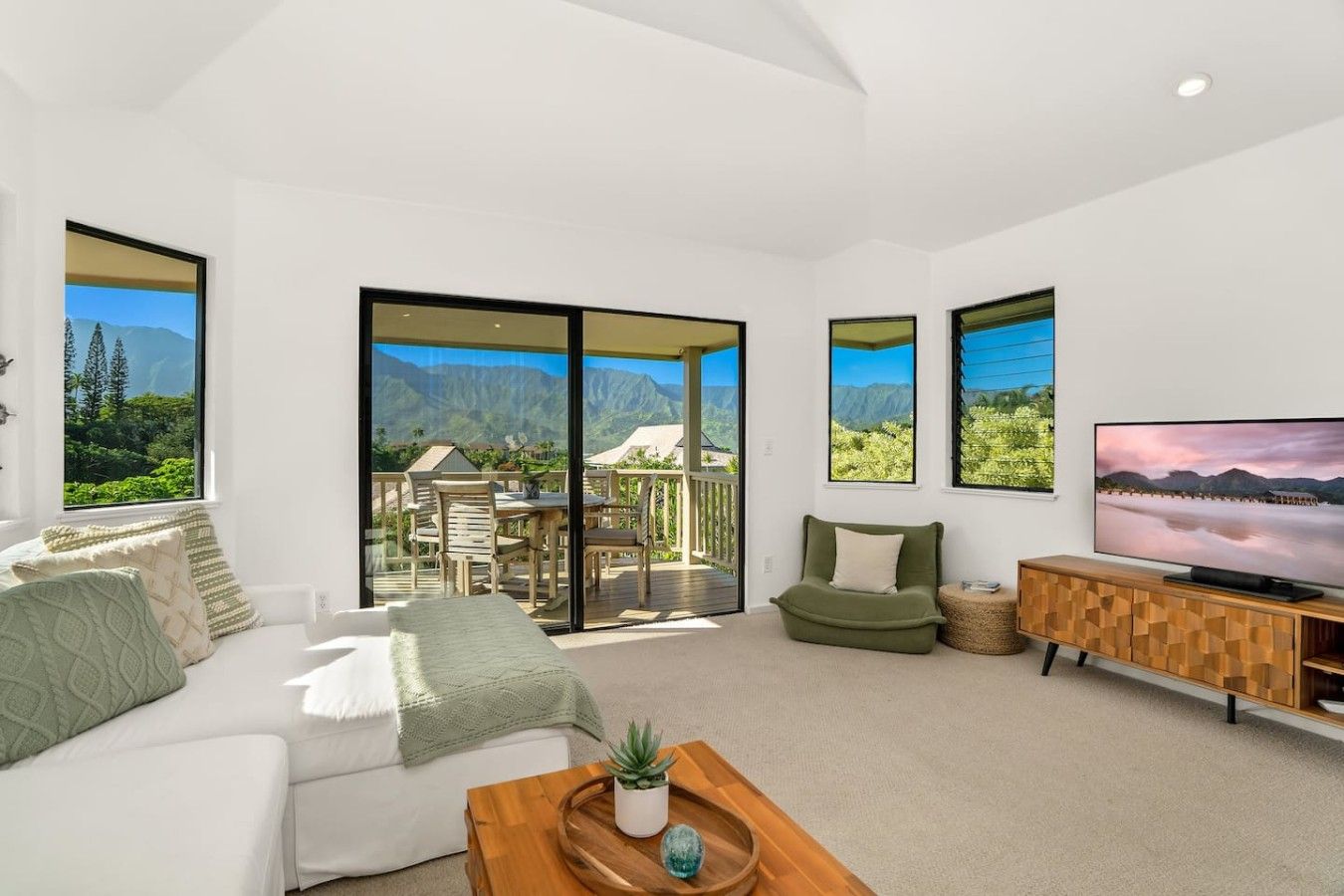 Ein modernes Wohnzimmer mit einer Couch, einem Couchtisch mit einer Pflanze, einem Fernseher und einem Balkon mit Bergblick.