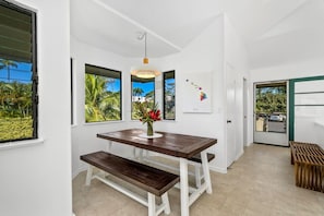 Un espace repas moderne avec une table et un banc en bois, de grandes fenêtres offrant une vue sur la végétation tropicale et un luminaire suspendu.