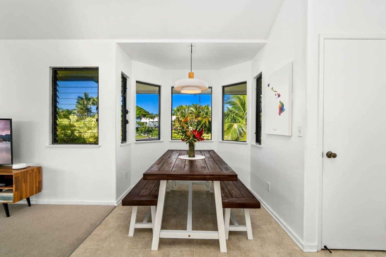 Un espace repas avec une table en bois, des murs blancs et de grandes fenêtres offrant une vue sur la végétation tropicale.