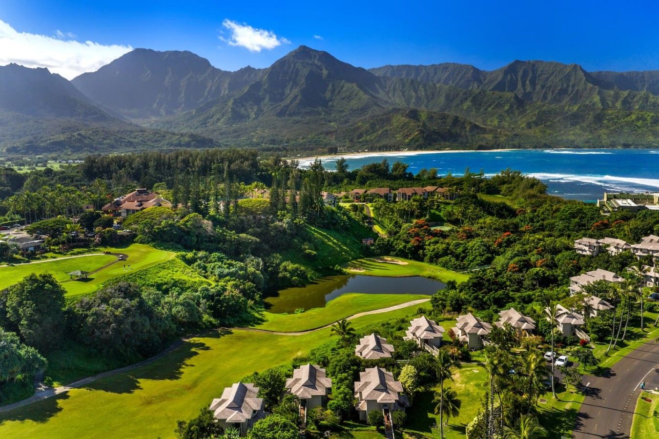 Un parcours de golf avec un étang, entouré d’une végétation luxuriante et dominé par une chaîne de montagnes en arrière-plan.