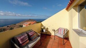 Uma varanda com sofá e poltrona, uma mesa pequena e vista para o mar.