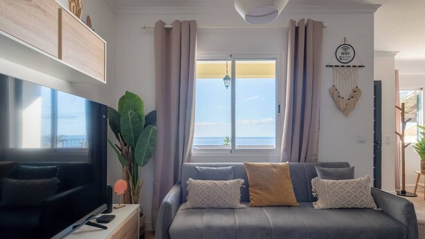 Uma sala de estar moderna com um sofá cinzento, uma televisão de tela plana e uma janela grande com vista para o mar.