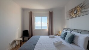 Quarto de hotel com cama, uma cadeira, janela com cortinas e uma obra de arte na parede.