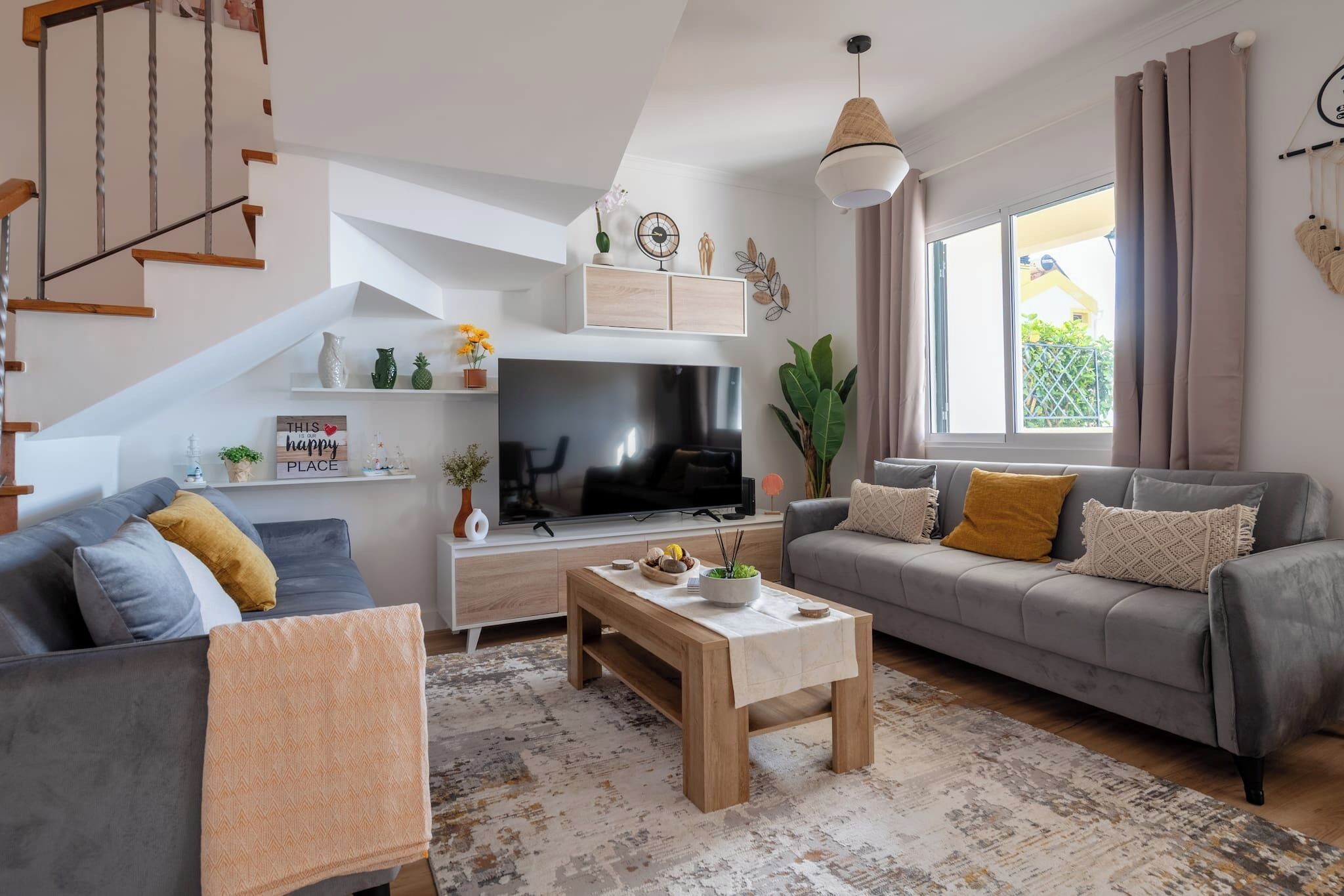 A modern living room with a staircase, a flat-screen TV, a sofa, a coffee table, and a large window.