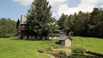 Une cabane en rondins avec une cheminée, entourée d’une pelouse bien entretenue et d’un petit étang.