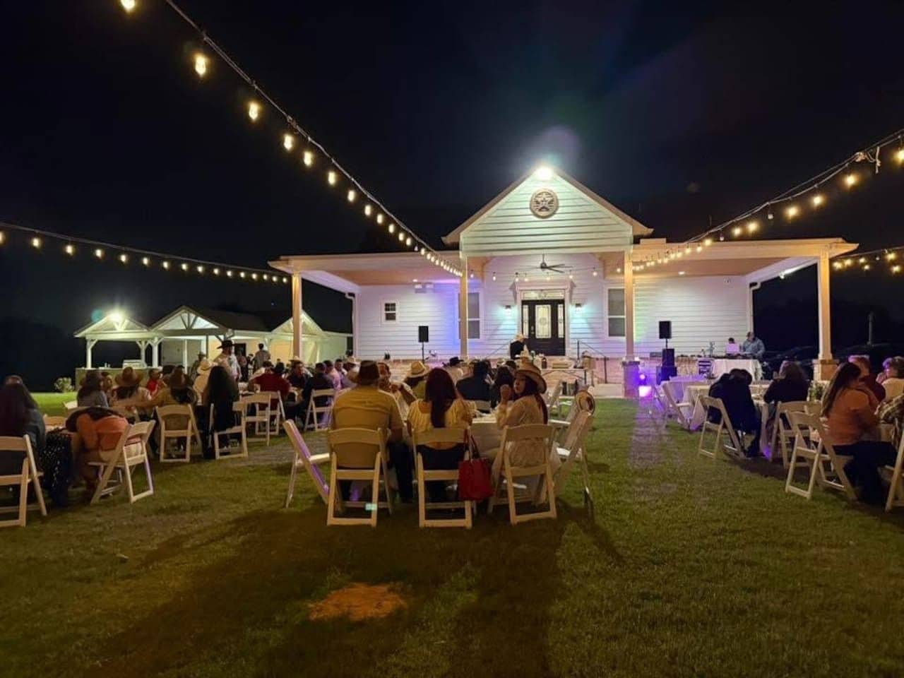 Un événement en plein air où les participants sont assis à des tables sous des guirlandes lumineuses, avec un bâtiment blanc doté d'un porche et une scène où une personne prend la parole.