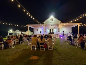 Evento ao ar livre com pessoas sentadas em mesas sob luzes pendentes, um prédio branco com varanda e um palco com uma pessoa falando.