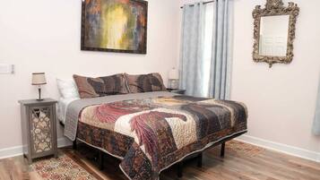 A bedroom with a bed, a nightstand, a lamp, a painting, a mirror, and a window with curtains.