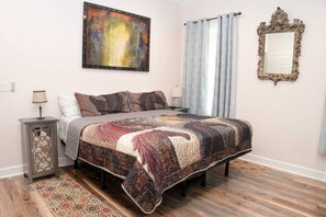 A bedroom with a bed, a nightstand, a lamp, a painting, a mirror, and a window with curtains.