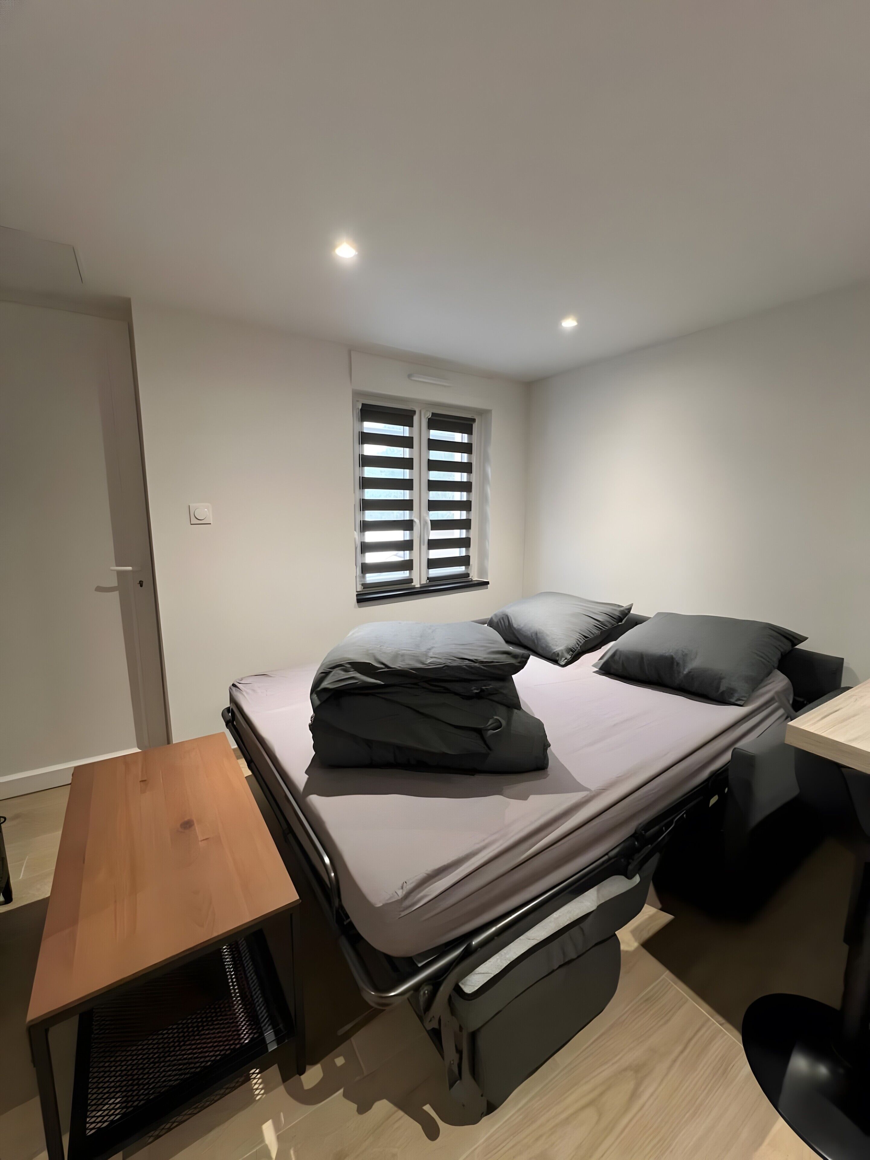 Ein kleines Schlafzimmer mit einem Klappbett, einem kleinen Holztisch und einem Fenster mit Jalousien.