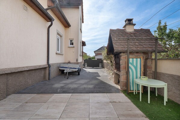 Un étroit sentier pavé menant à une entrée fermée par un portail, une petite table avec un panier, et un bâtiment doté d’un toit en tuiles et d’une fenêtre.