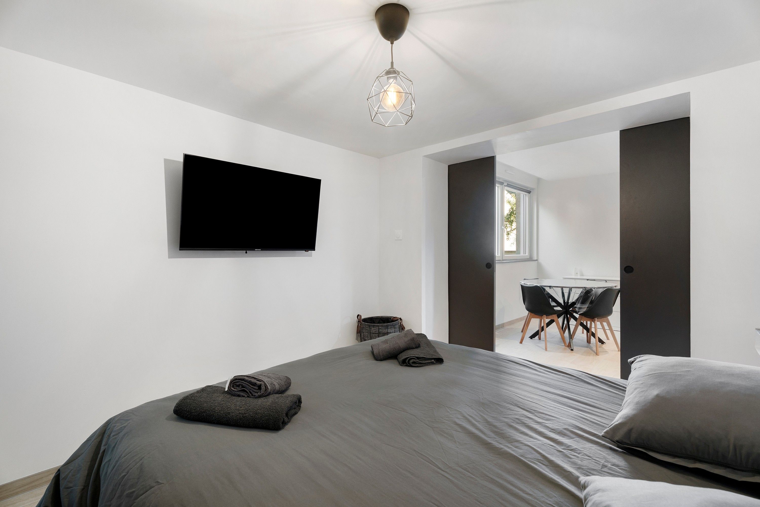 Ein modernes Schlafzimmer mit einem großen Bett, einem flachbildfernseher an der Wand und einem Essbereich mit Tisch und Stühlen, der durch eine offene Tür sichtbar ist.