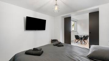 Ein modernes Schlafzimmer mit einem großen Bett, einem flachbildfernseher an der Wand und einem Essbereich mit Tisch und Stühlen, der durch eine offene Tür sichtbar ist.