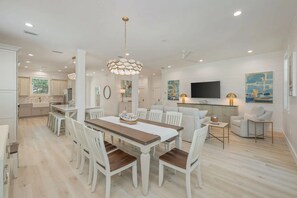 A modern kitchen with white cabinets, a dining area with a wooden table and white chairs, and a living area with a sofa and a TV. - Beacon: 30A beaches, sleeps 26, private pool! (Santa Rosa Beach)