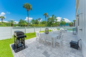 An outdoor patio with a grill, white chairs, and a table, surrounded by a pool and greenery. - Beacon: 30A beaches, sleeps 26, private pool! (Santa Rosa Beach)