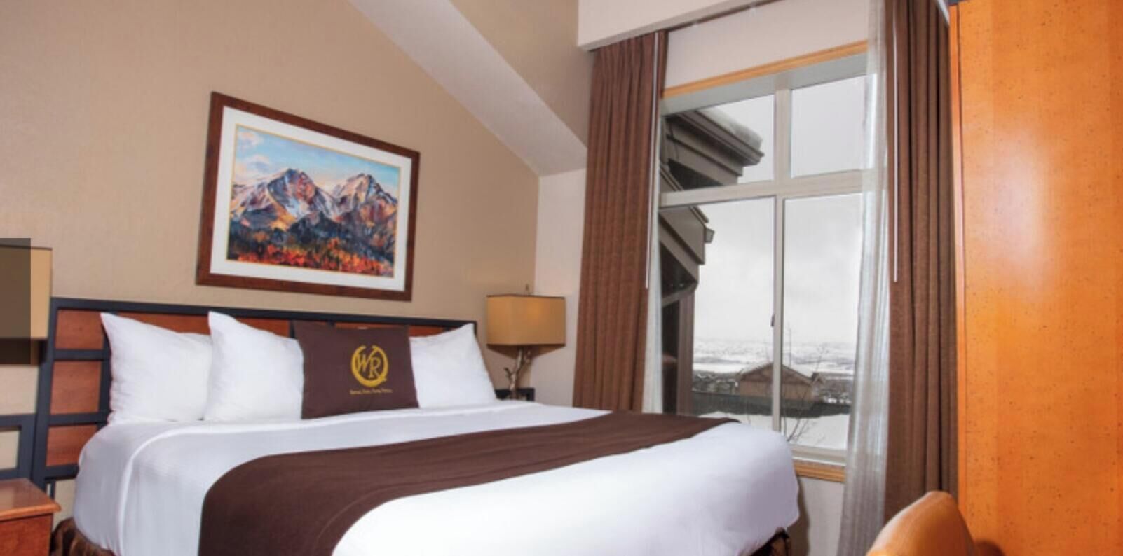 Une chambre d’hôtel avec un lit, un tableau représentant des montagnes, une fenêtre avec des rideaux et une lampe.