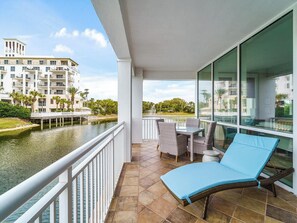 A balcony with a view of a water body and a building, furnished with a table, chairs, and a lounge chair. - Kiva Village A 105 - Golf Course Views & Luxury Beach Club Access at Kiva Village (Gulf Shores)