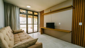 A living room with a sofa, a television on a wooden shelf, and a sliding glass door leading to an outdoor area. - Resort Apartment with Access to Coqueiro Beach (Luís Correia)