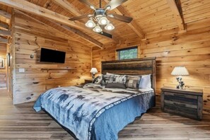 Ein Schlafzimmer einer Holzhütte mit einem Bett, zwei Nachttischen, einem Deckenventilator und einem an der Wand montierten Fernseher.