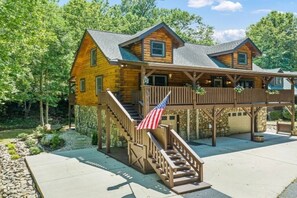 A two-story log cabin with a covered porch and a flag. - Breezy Creek Retreat-Spectacular Waterfront Cabin! (Maggie Valley)