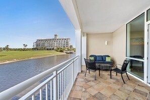 A balcony with a view of a river, a building across the water, and a seating area with chairs and a sofa. - Kiva Village A 101 - Golf Course & Sunset Views Luxury 3BR Condo Near Fort Morgan Beach (Gulf Shores)