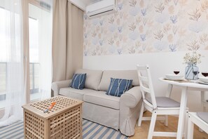Una sala de estar con un sofá, una mesa de comedor con sillas, una mesita de madera y una ventana con cortinas translúcidas.